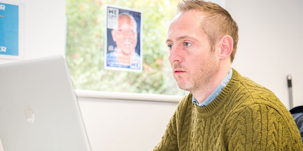 A person in an olive cable-knit jumper looking at a laptop screen in an office, with an "It Starts With Me" poster behind.