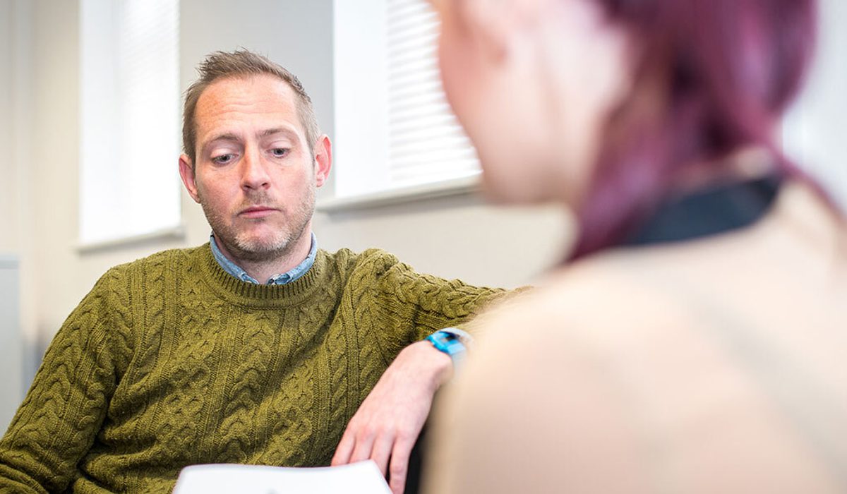 A person in an olive cable-knit jumper listening attentively to another person seen from behind, in an office.