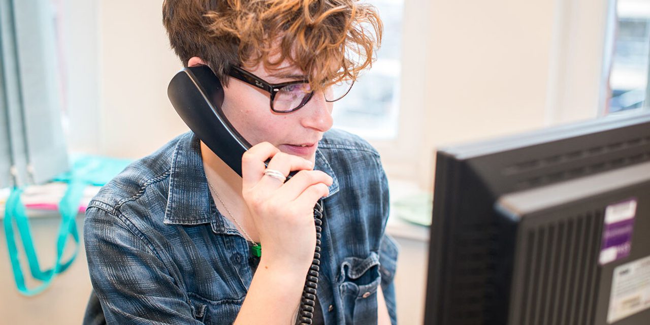 A person with curly hair and glasses, in a denim shirt, talking on a desk phone while looking at a computer monitor.
