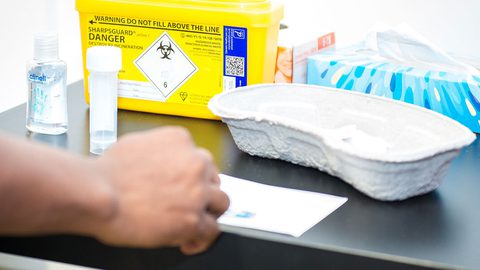 A hand on a black table beside a yellow clinical sharps bin marked "Danger", hand sanitiser, a sample tube and a tissue box.