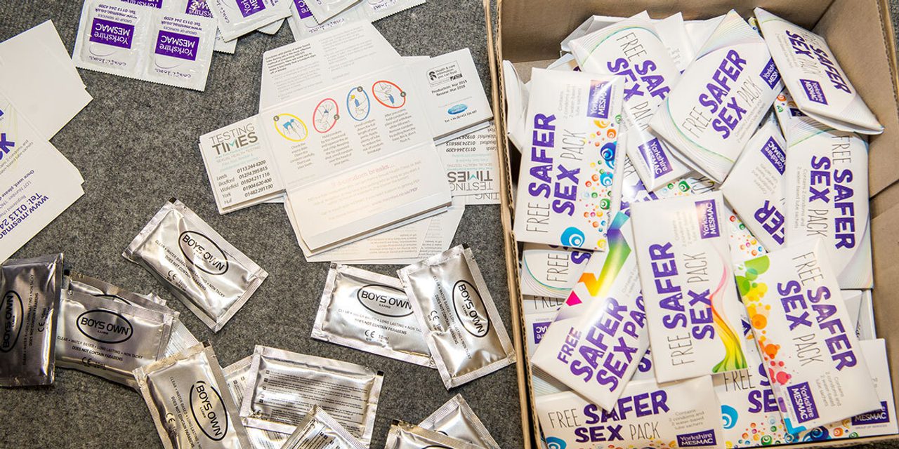 Loose condom sachets and "Testing Times" leaflets beside an open box of "Free Safer Sex Pack" boxes on grey carpet.