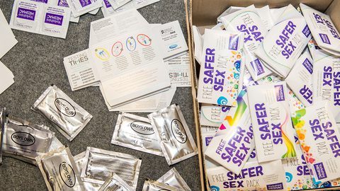 Loose condom sachets and "Testing Times" leaflets beside an open box of "Free Safer Sex Pack" boxes on grey carpet.