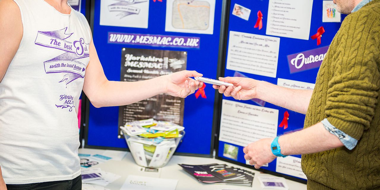 Close-up of hands exchanging a condom packet over an information stall with a Yorkshire MESMAC display board and red ribbons; one person in a "The best sex with the least harm" vest.
