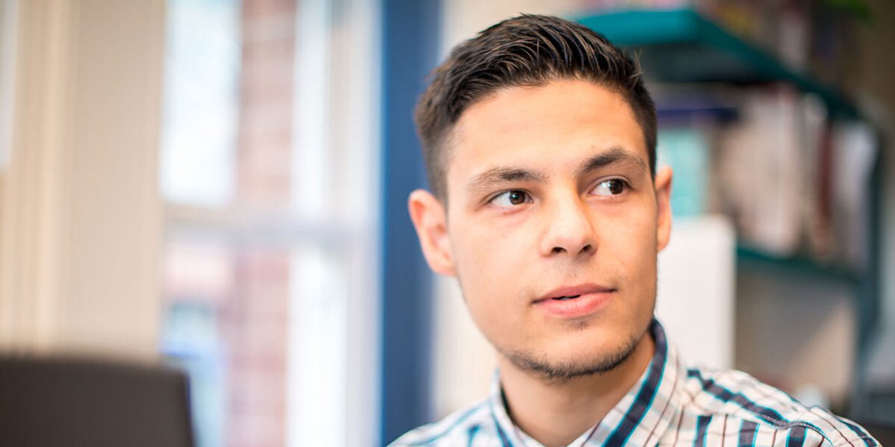 Close-up portrait of a person with short hair and light stubble in a checked shirt, looking to the side in an office.