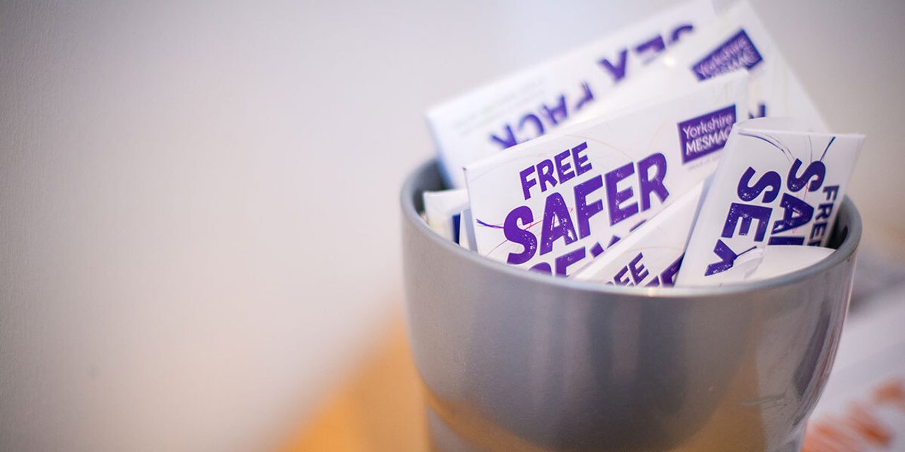 Close-up of a metal cup filled with Yorkshire MESMAC "Free Safer Sex" packs.