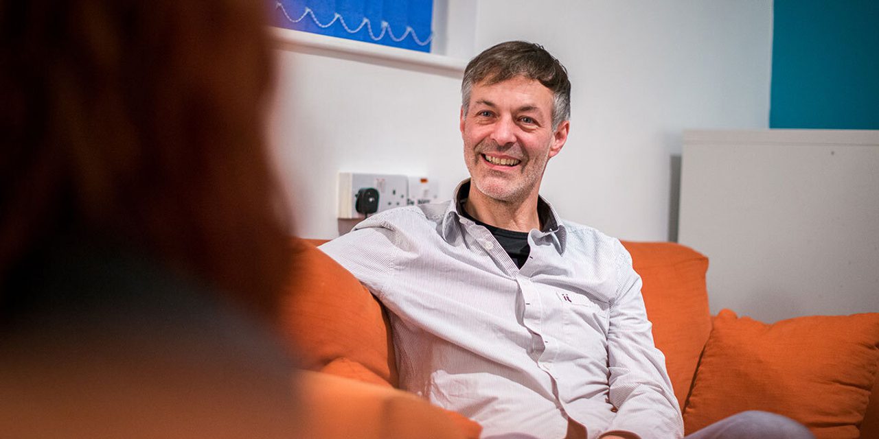 A person with greying hair in a striped shirt smiling while sitting on an orange sofa, with another person seen from behind.