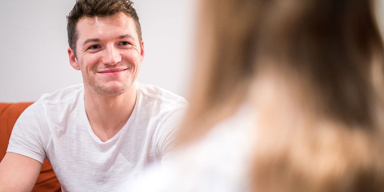 A person in a white t-shirt smiling while sitting and talking to another person seen from behind.