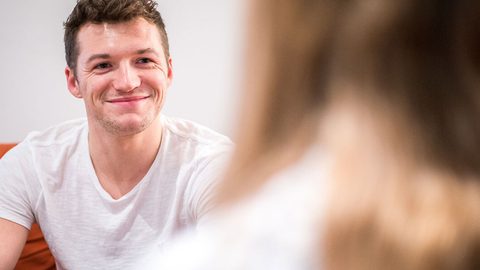 A person in a white t-shirt smiling while sitting and talking to another person seen from behind.