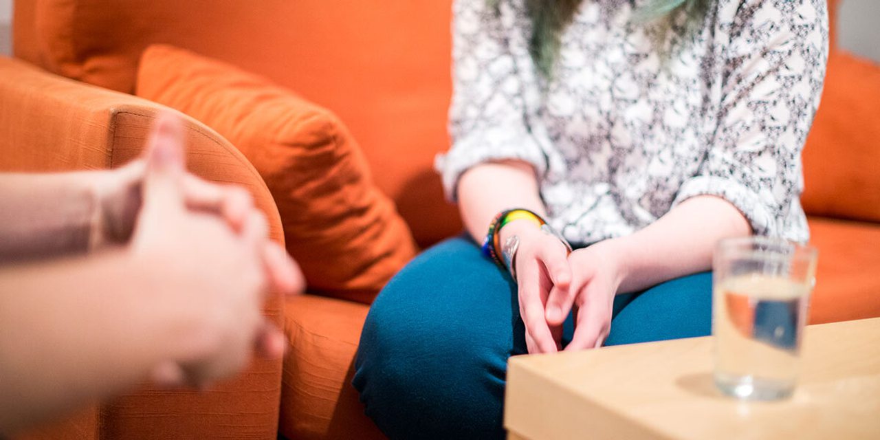 Close-up of a person in a patterned top with teal-tipped hair sitting on an orange sofa, hands on their lap, a glass of water on a side table.