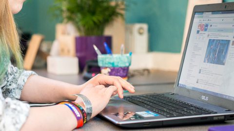 A person with teal-tipped hair and rainbow wristbands typing on a laptop showing a social media feed.