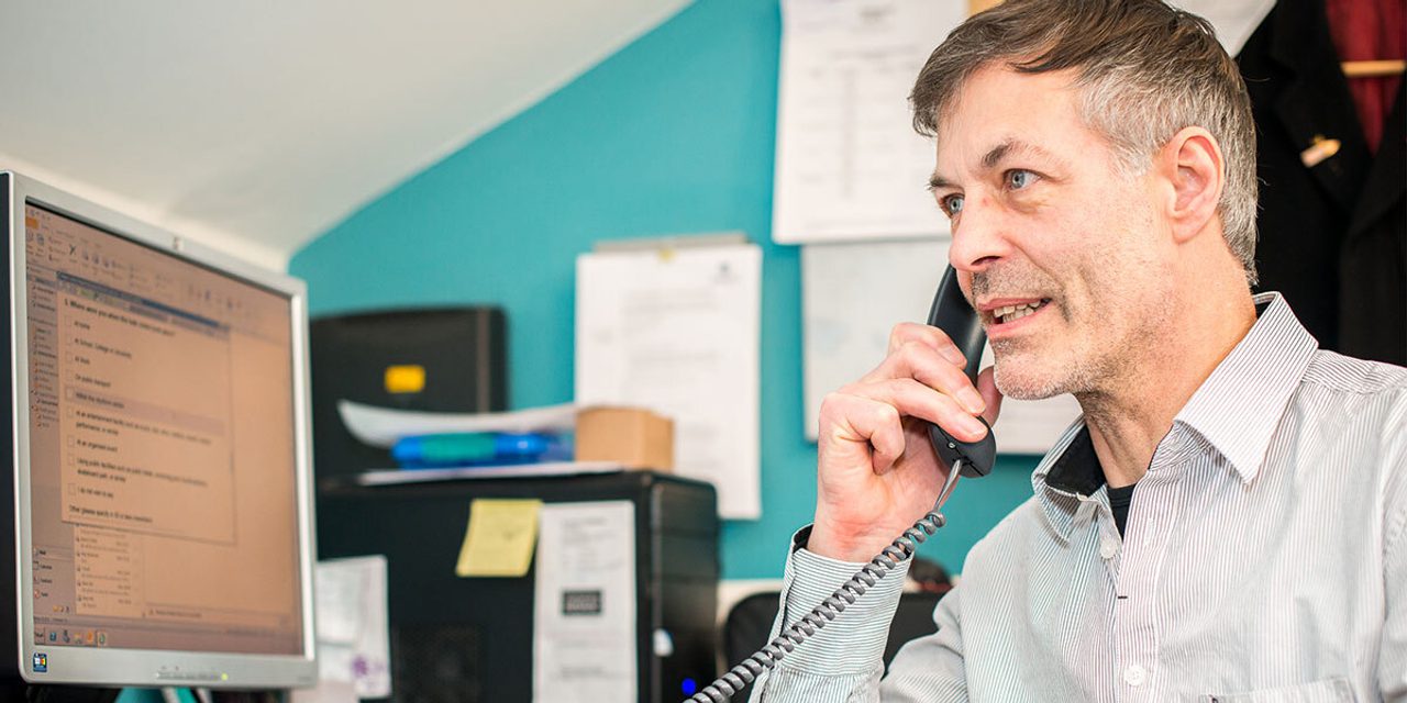 A person with greying hair in a striped shirt talking on a desk phone at a computer in a teal-walled office.