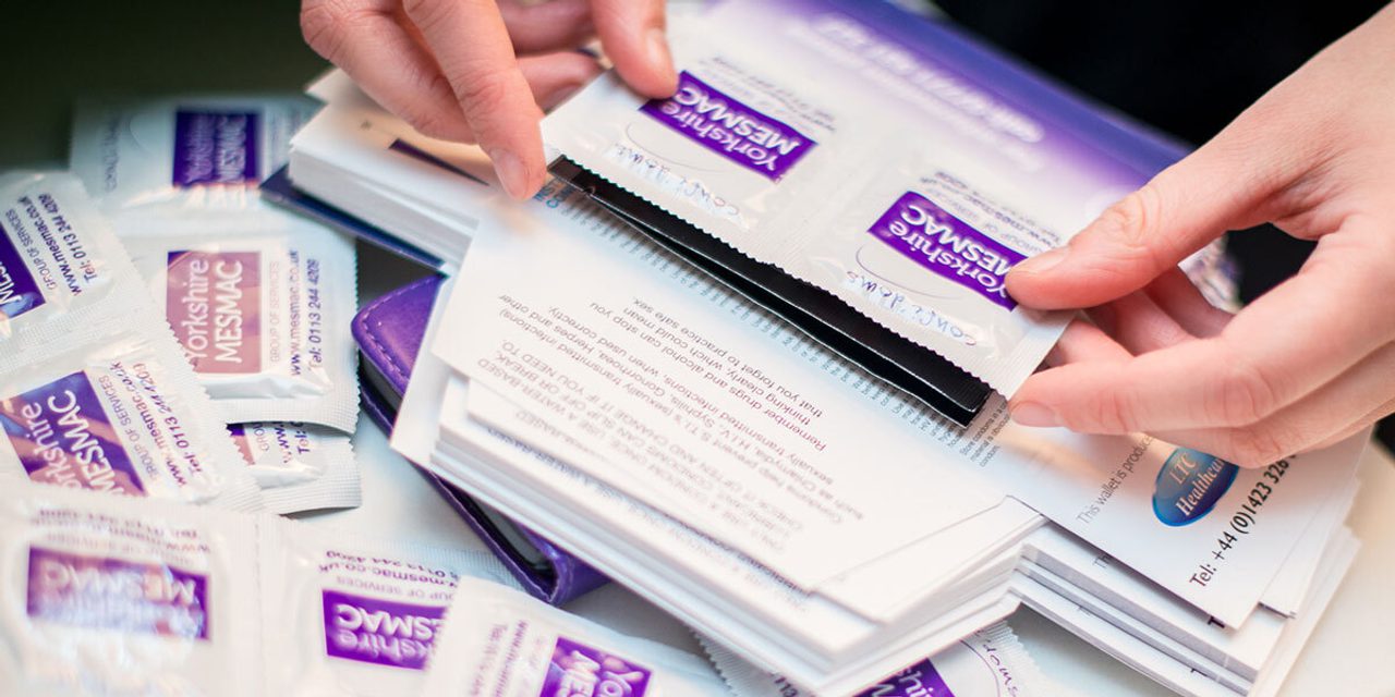 Close-up of hands sorting through a pile of Yorkshire MESMAC condom packets on a table.