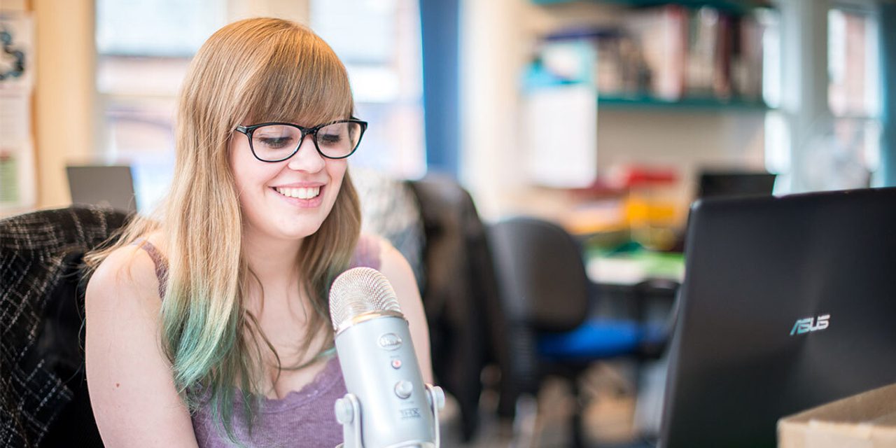 A person with long hair and glasses smiling while speaking into a microphone at a desk, with a laptop nearby.