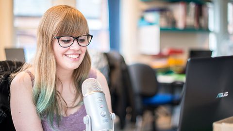 A person with long hair and glasses smiling while speaking into a microphone at a desk, with a laptop nearby.
