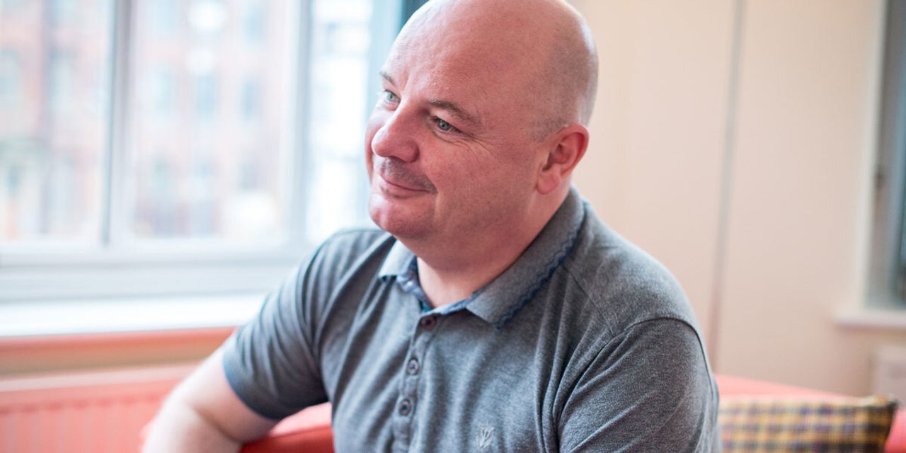Portrait of a person with a shaved head in a grey polo shirt, smiling softly while sitting on an orange sofa by a window.