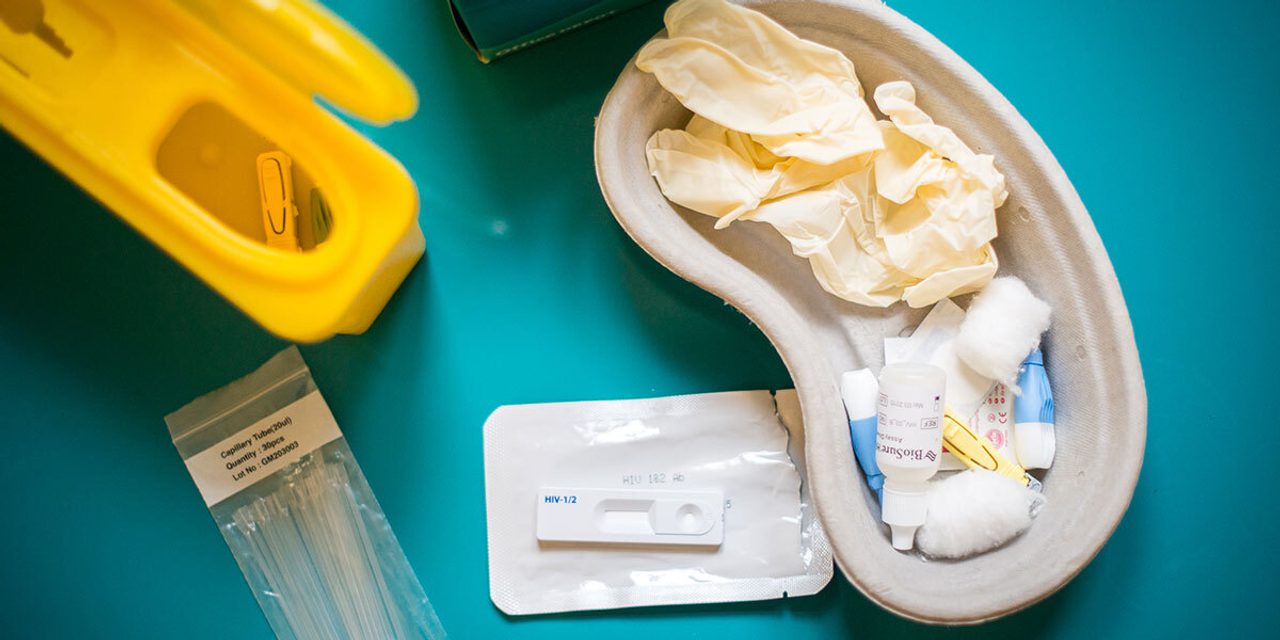 An HIV self-test kit laid out on a teal surface: a yellow sharps bin, capillary tubes, a kidney dish with gloves and swabs, and a BioSure test cassette.