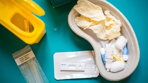 An HIV self-test kit laid out on a teal surface: a yellow sharps bin, capillary tubes, a kidney dish with gloves and swabs, and a BioSure test cassette.