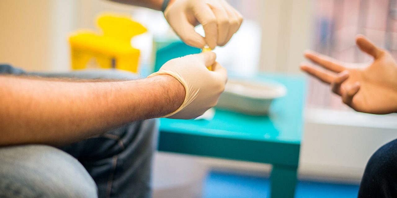 Close-up of gloved hands preparing a finger-prick test, with another person's hand gesturing opposite.