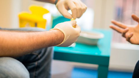 Close-up of gloved hands preparing a finger-prick test, with another person's hand gesturing opposite.