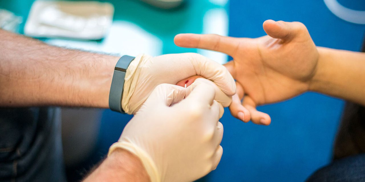 Close-up of gloved hands performing a finger-prick blood test on another person's finger, a fitness band on one wrist.