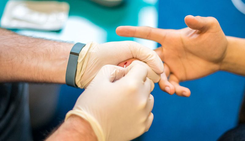 Close-up of gloved hands performing a finger-prick blood test on another person's finger, a fitness band on one wrist.