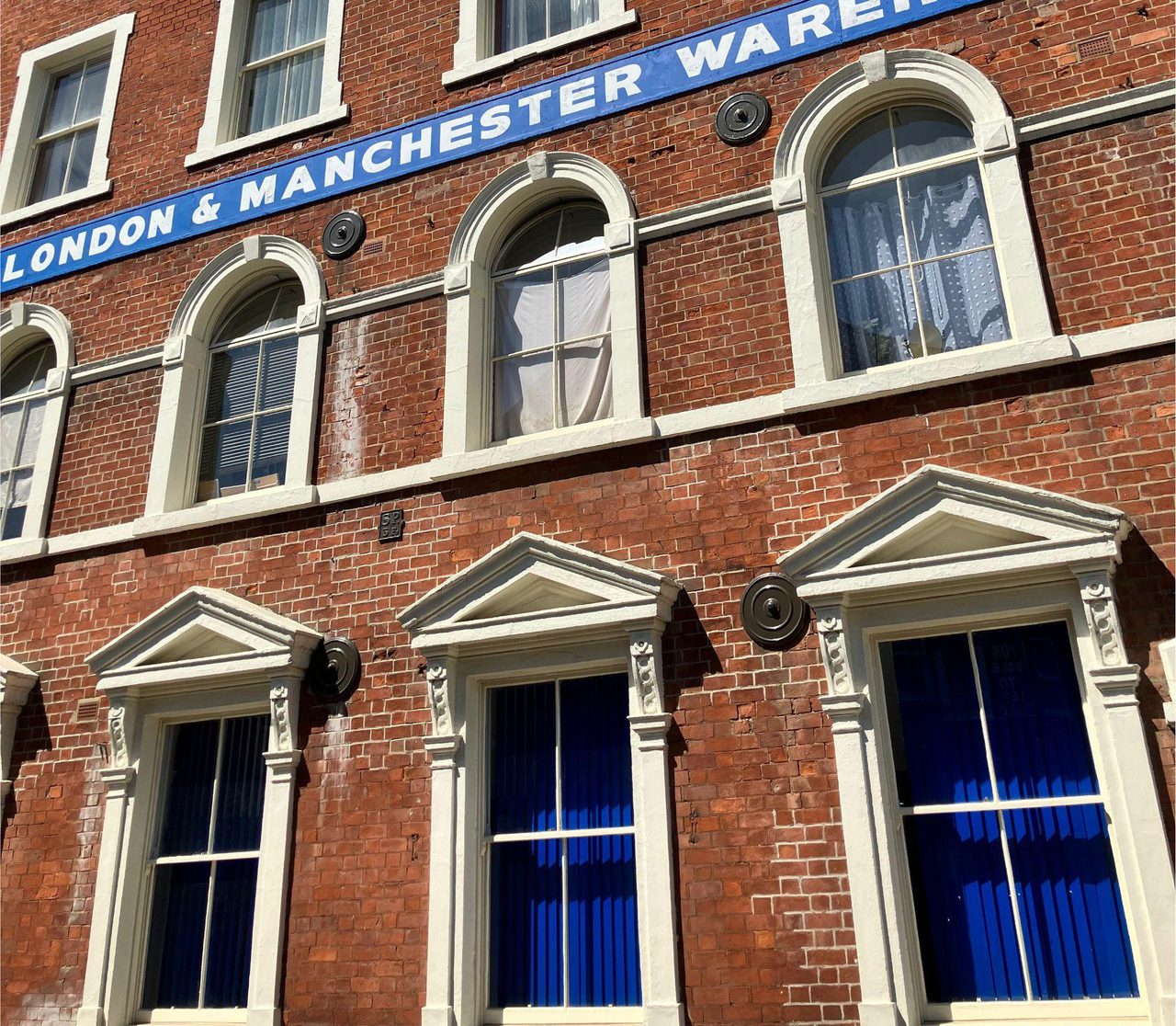 Red brick warehouse facade with arched sash windows and a blue sign reading "London & Manchester Warehouse", under a clear blue sky.