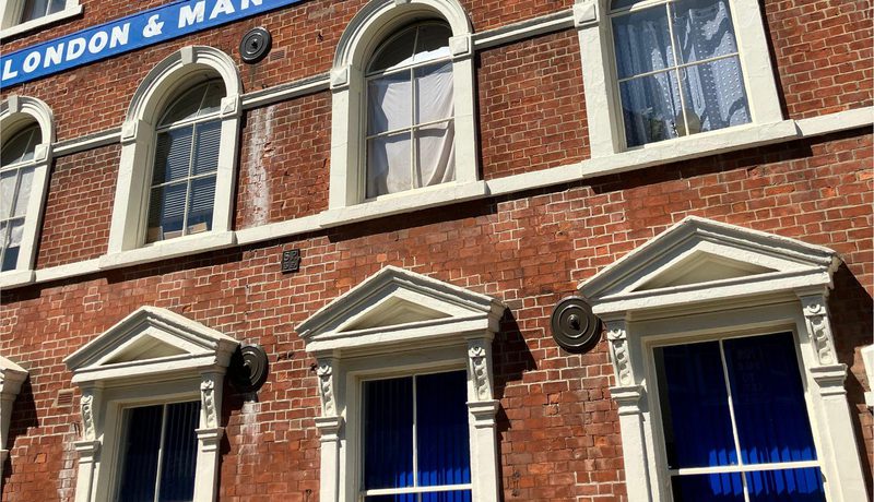 Red brick warehouse facade with arched sash windows and a blue sign reading "London & Manchester Warehouse", under a clear blue sky.