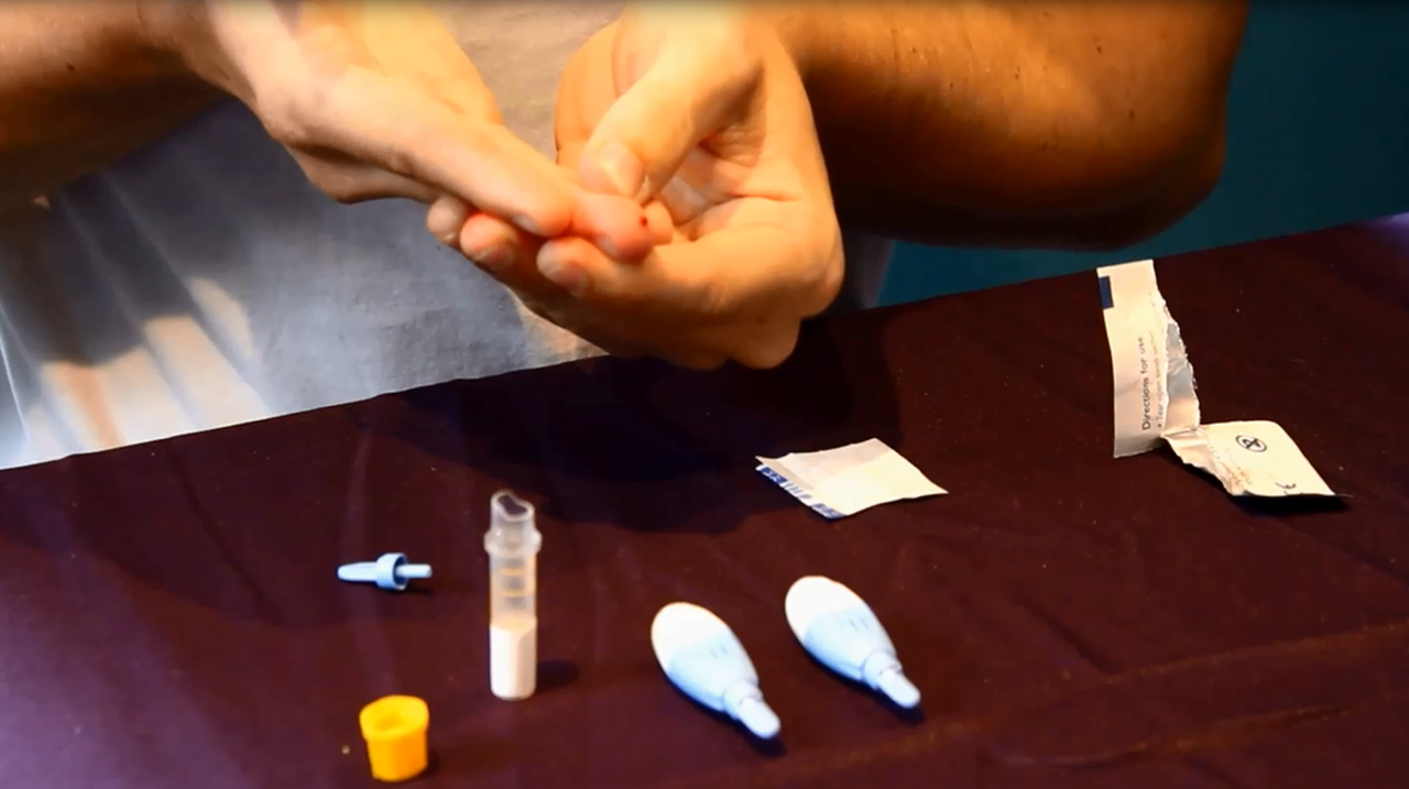 Close-up of hands preparing a home test sampling kit on a dark red surface, with a sample tube, dropper pipettes and instructions.