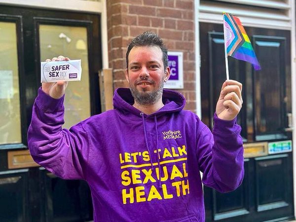 A person in a purple Yorkshire MESMAC "Let's Talk Sexual Health" hoodie holding a safer-sex pack and a Progress Pride flag in a street.