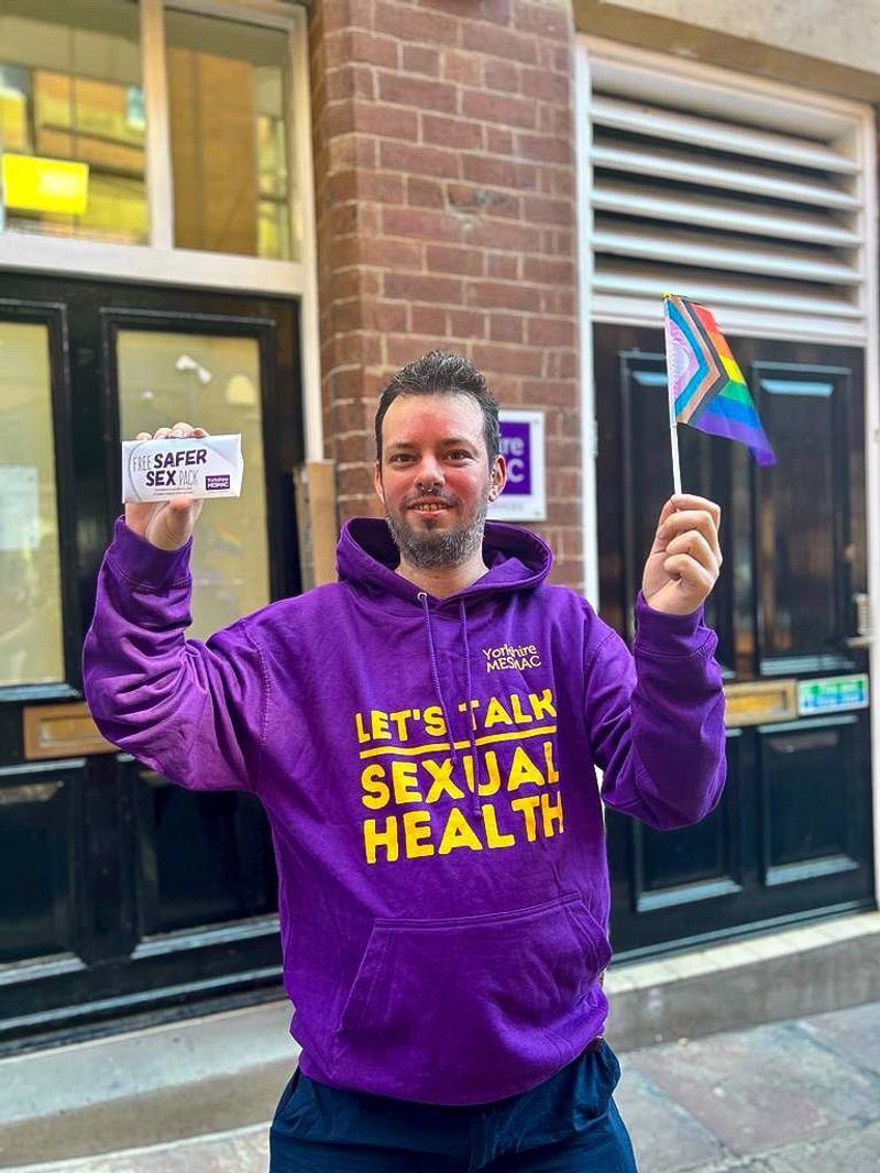 A person in a purple Yorkshire MESMAC "Let's Talk Sexual Health" hoodie holding a safer-sex pack and a Progress Pride flag in a street.