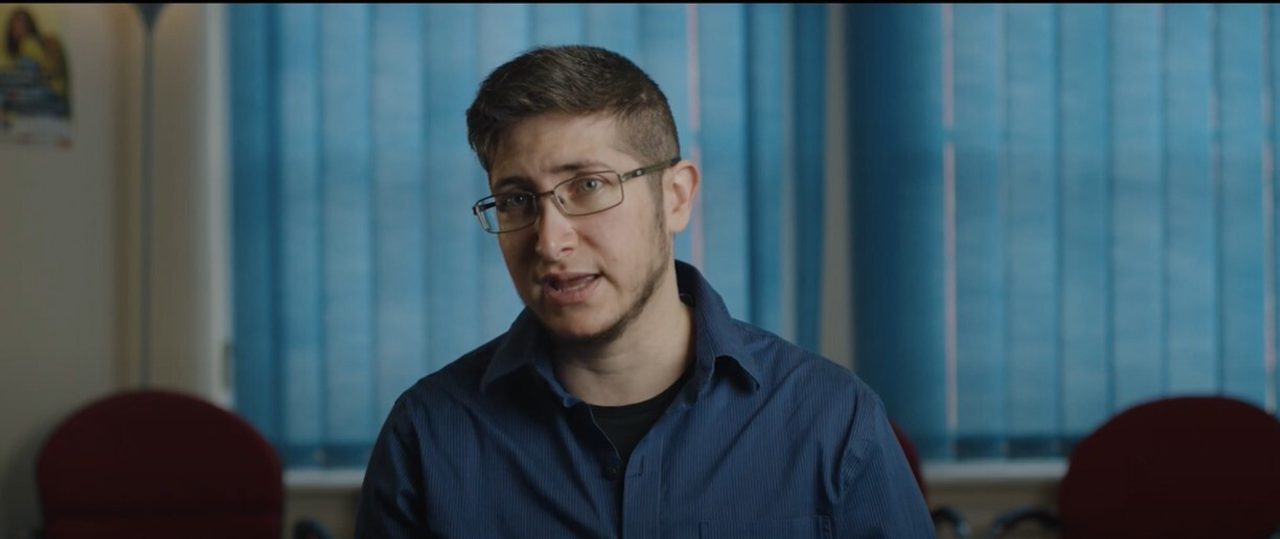 A person in glasses and a blue shirt talking, seated in an office (a video still).