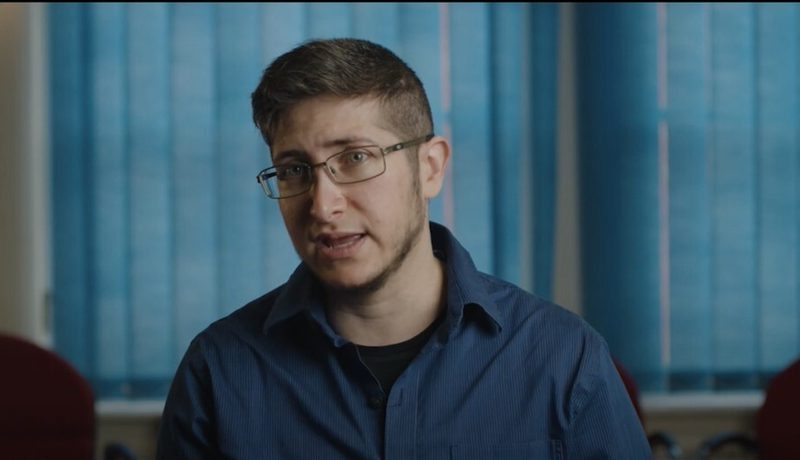 A person in glasses and a blue shirt talking, seated in an office (a video still).